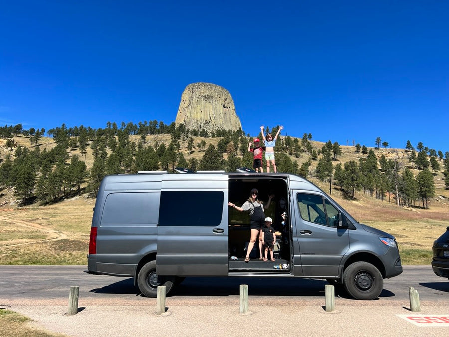 A family poses with their RV rental at Devils Tower. Kids wave from the roof, adults and a child are in the open side door.