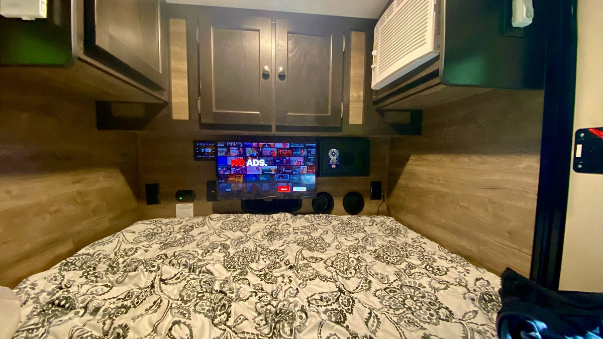 Cozy RV rental bedroom features a mounted TV for entertainment, patterned bed, overhead cabinets, and an AC unit. — photo 11