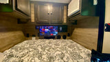 Cozy RV rental bedroom features a mounted TV for entertainment, patterned bed, overhead cabinets, and an AC unit.