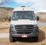 Front view of an RV rental: a white Mercedes Sprinter camper van parked by a lake, with hills and trees in the background.