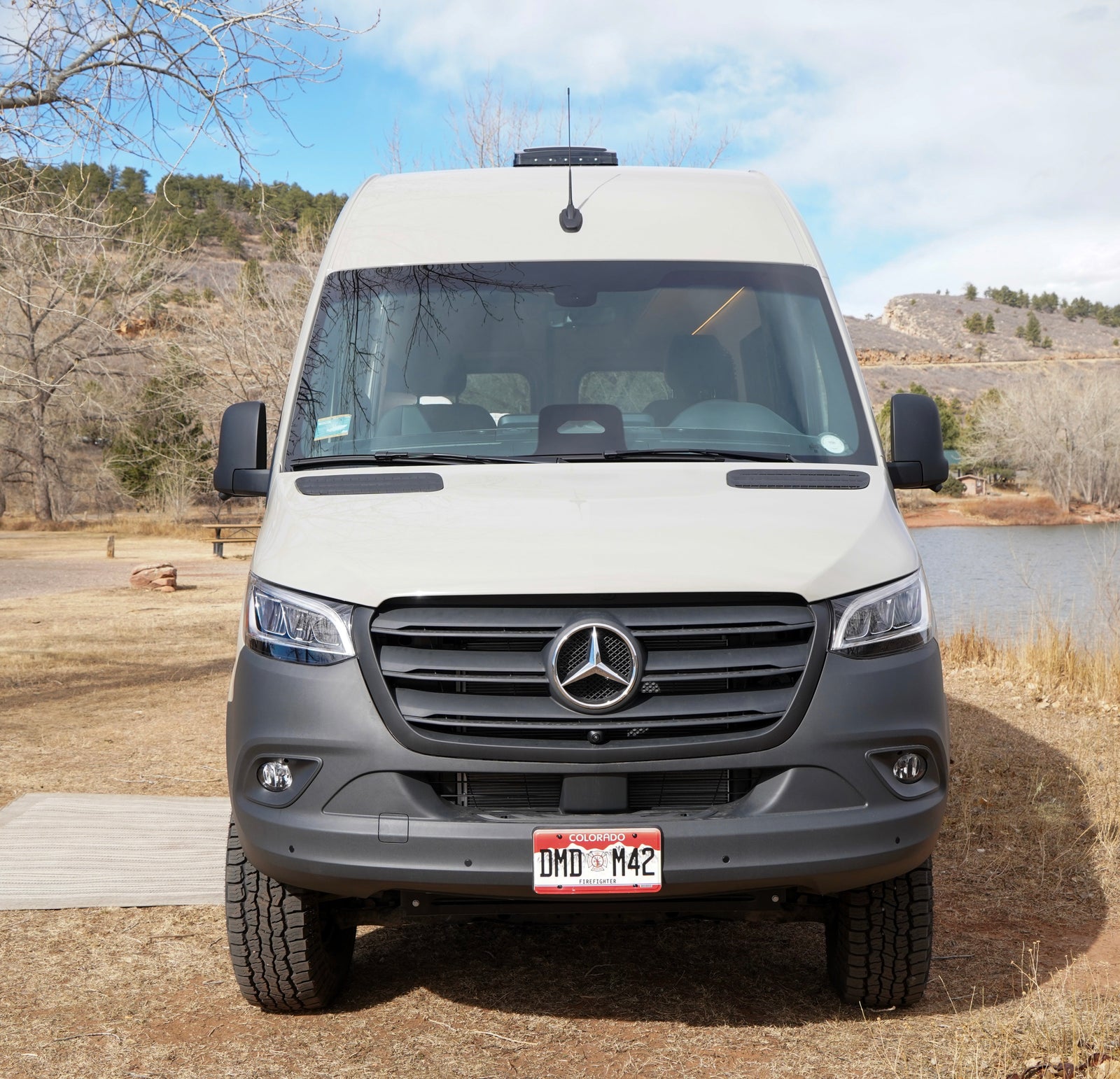 Front view of an RV rental: a white Mercedes Sprinter camper van parked by a lake, with hills and trees in the background.