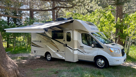 A tan Compass rv rental with an extended awning is parked in a sunny, wooded campsite, ready for your next adventure.