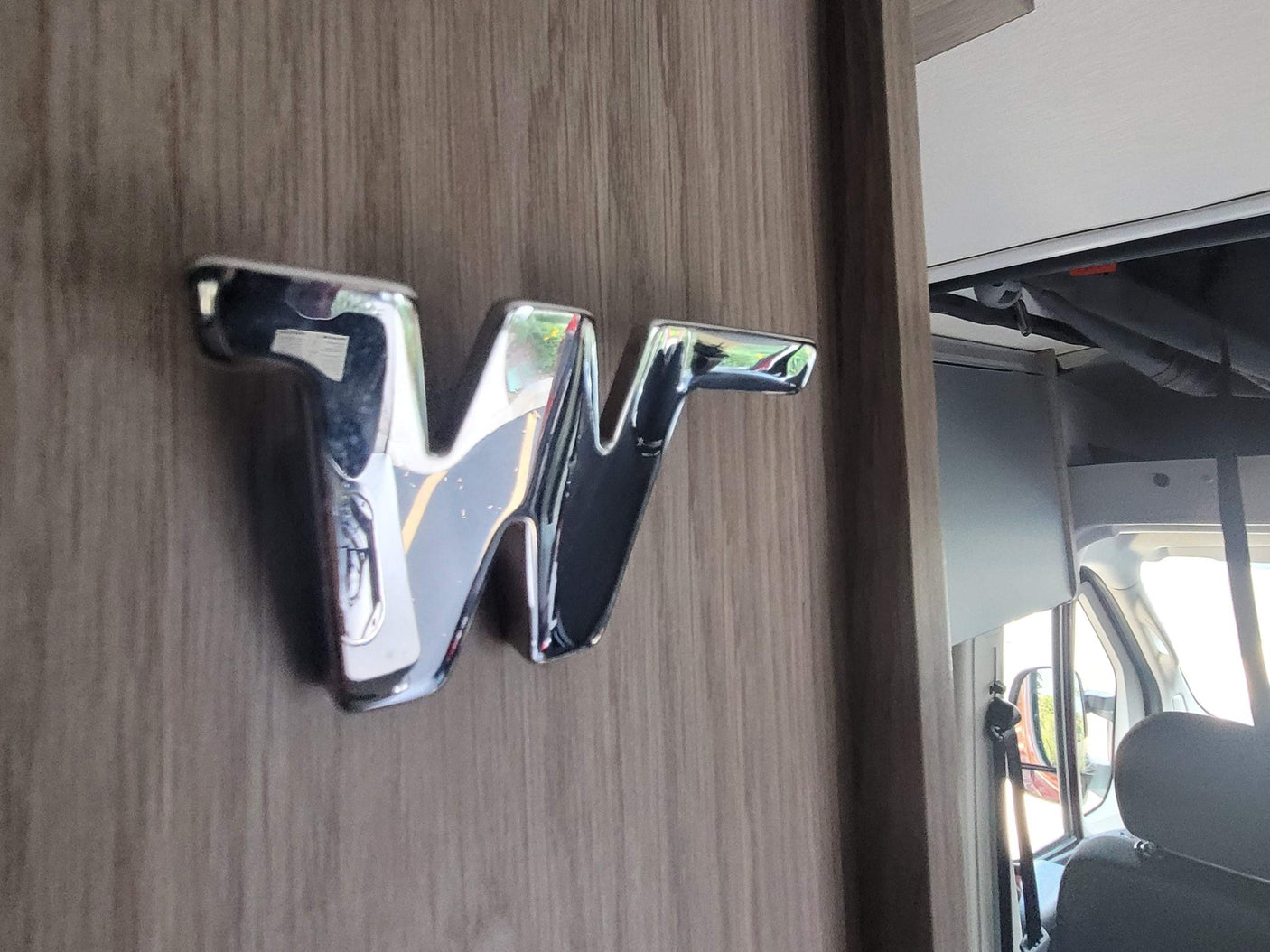 Winnebago RV rental interior with a chrome 'W' logo on wood paneling, reflecting the road and cabin details.