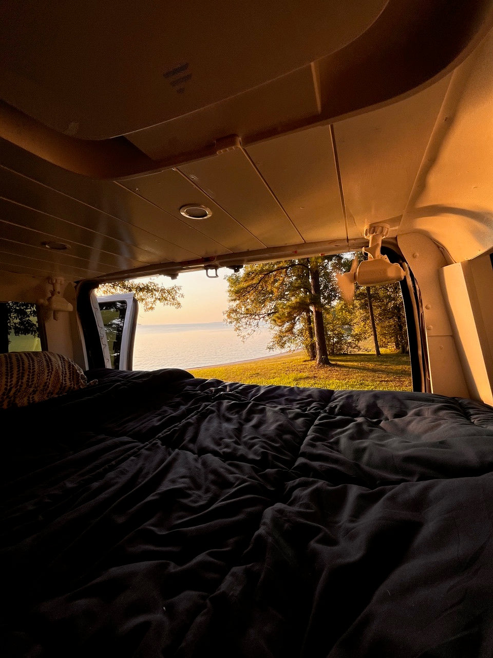 Cozy RV rental view: a comfy bed opens to a serene lakeside landscape at sunrise. Perfect for van life adventures.