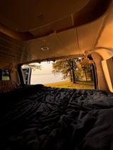 Cozy RV rental view: a comfy bed opens to a serene lakeside landscape at sunrise. Perfect for van life adventures.