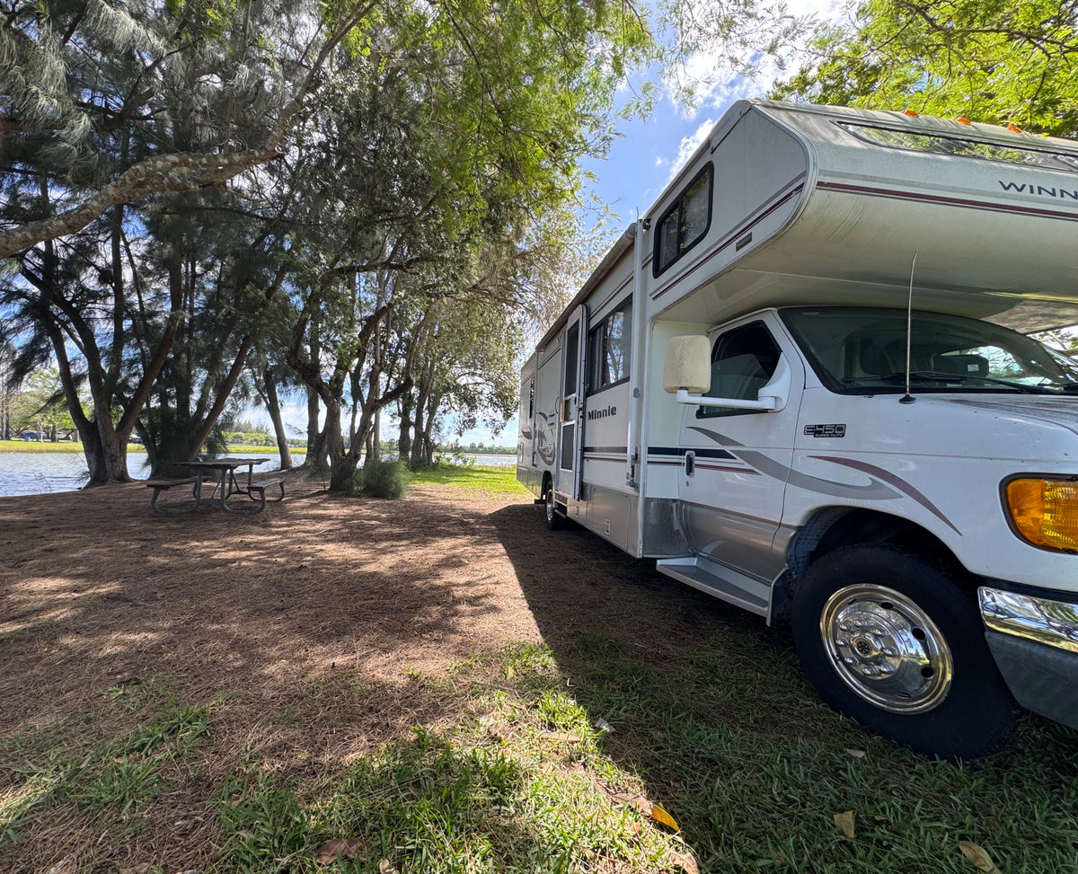 Winnebago Minnie RV rental by a tranquil lake, shaded by trees with a picnic table. Ideal for an outdoor escape.