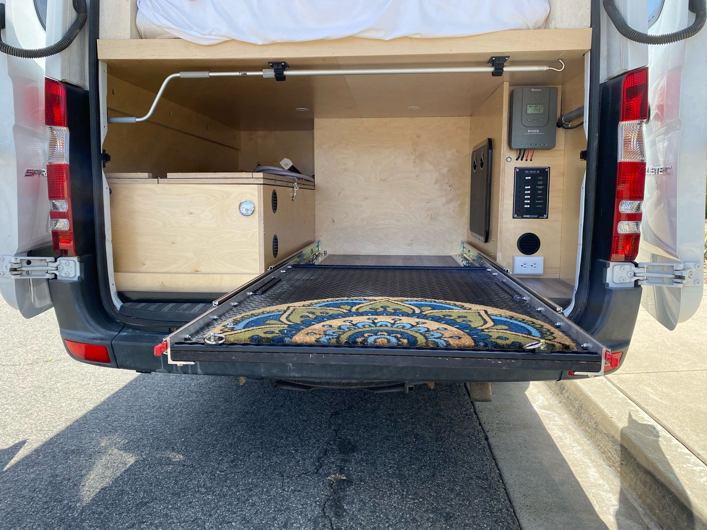 RV rental camper van interior with rear doors open, showing custom bed, pull-out storage, and power panel.