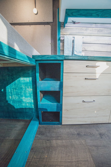 Compact RV rental interior featuring a turquoise bed frame, shelving, and wood drawers for smart storage.