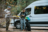 Two people and their dog set up for fly fishing from a white RV rental camper van in the woods.