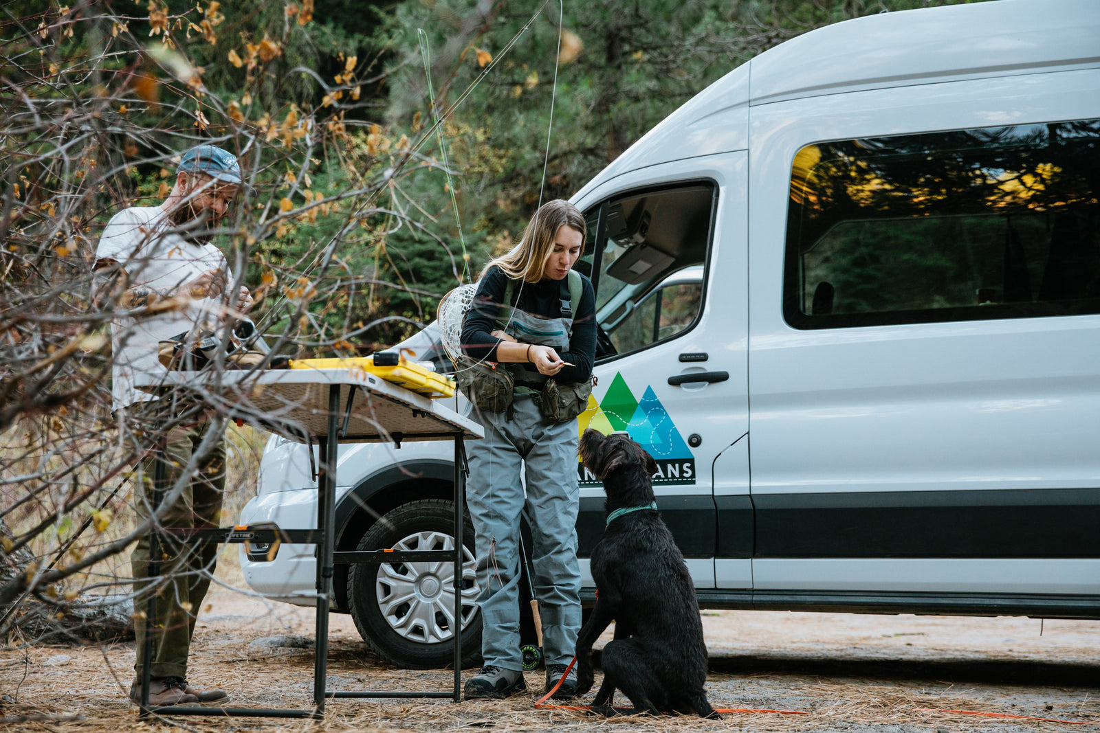 Two people and their dog set up for fly fishing from a white RV rental camper van in the woods.