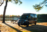 Black camper van RV rental parked lakeside, featuring a picnic table and trees. Enjoy serene nature and outdoor adventures under sunny skies.