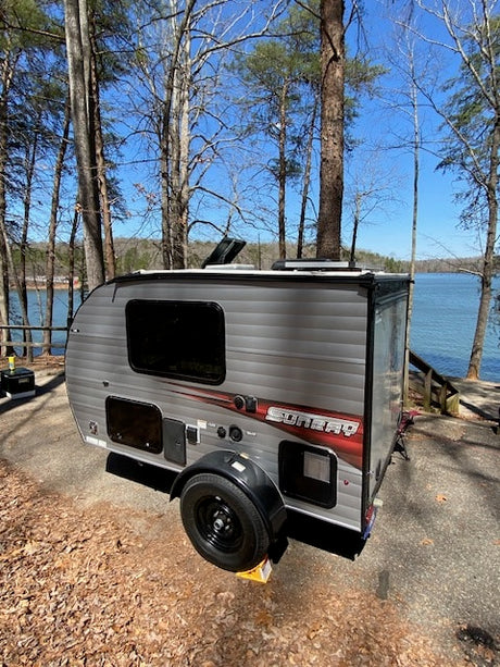 Sunny day RV rental at a scenic lakeside campsite. A gray travel trailer sits among trees by a blue lake, ideal for camping.