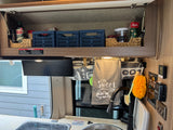 RV rental kitchen with organized shelf storage, sink, and controls, featuring a "Camper Sweet Camper" towel.