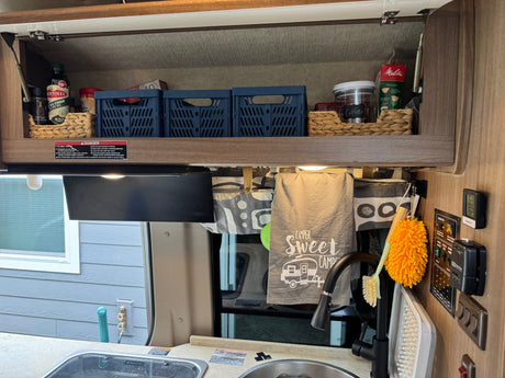 RV rental kitchen with organized shelf storage, sink, and controls, featuring a "Camper Sweet Camper" towel.