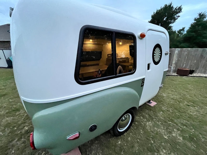 Cozy white & green retro camper, an RV rental, parked on grass. Interior lights glow, showing a comfy small trailer space. — photo 8