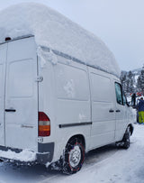 Heavy snow blankets this white Mercedes Sprinter van, ready for a winter RV rental adventure with tire chains.