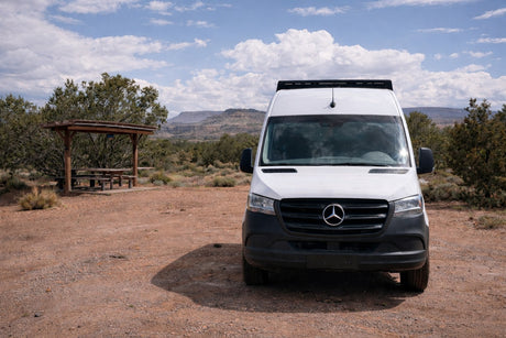 A white Mercedes Sprinter RV rental van in a rustic desert picnic area with a wooden shelter, under a vast cloudy sky.