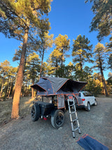 A gray rooftop tent on a utility trailer, with a ladder, is set up next to a white SUV in a sunny forest. Great for rv rental adventures.