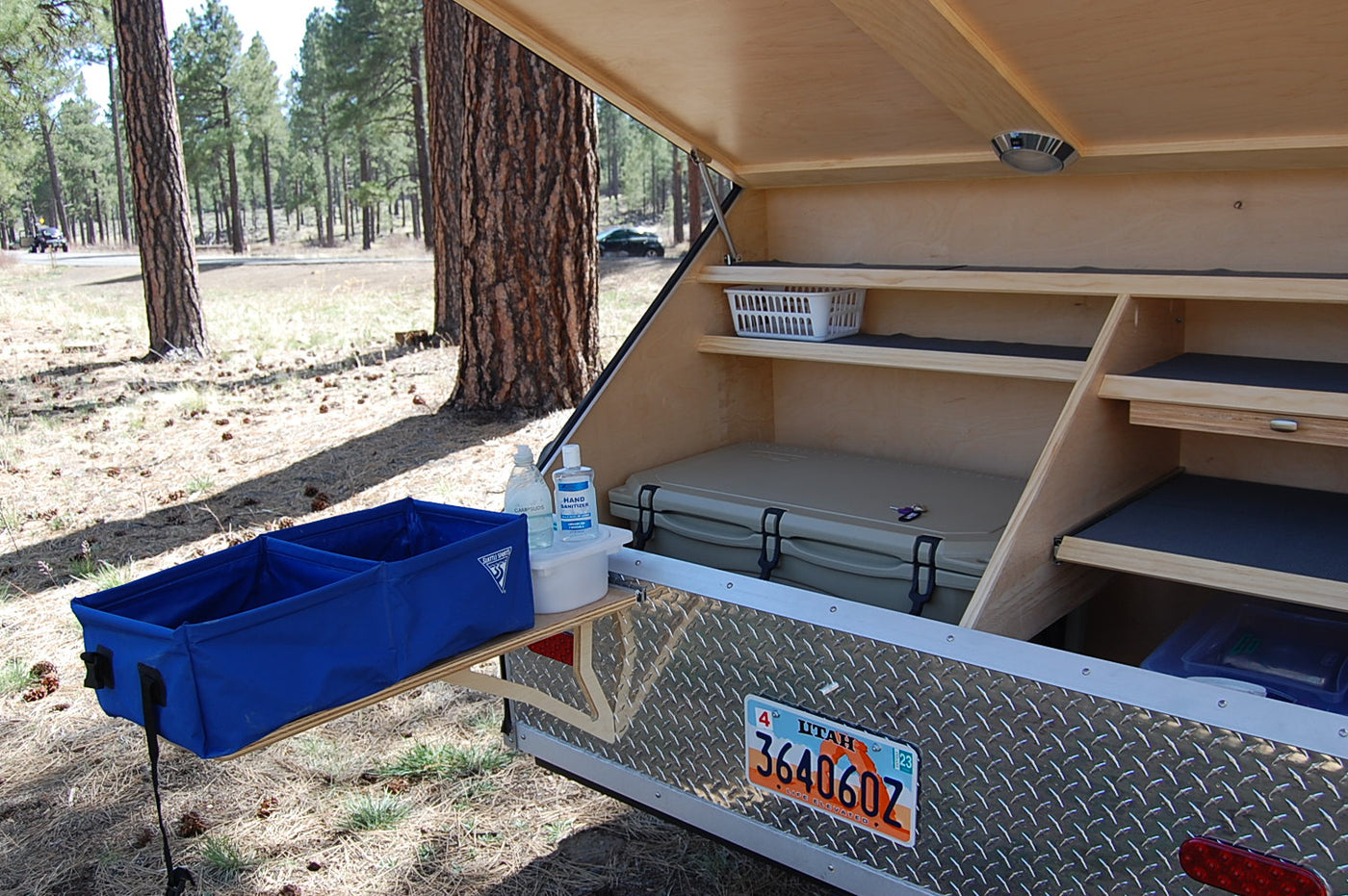 An RV rental's rear galley is open at a forest campsite, revealing shelves, a cooler, and an outdoor wash basin. — photo 6
