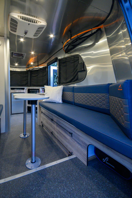 Modern rv rental interior: blue seating with storage, small table, polished aluminum walls, AC, and speckled floor.