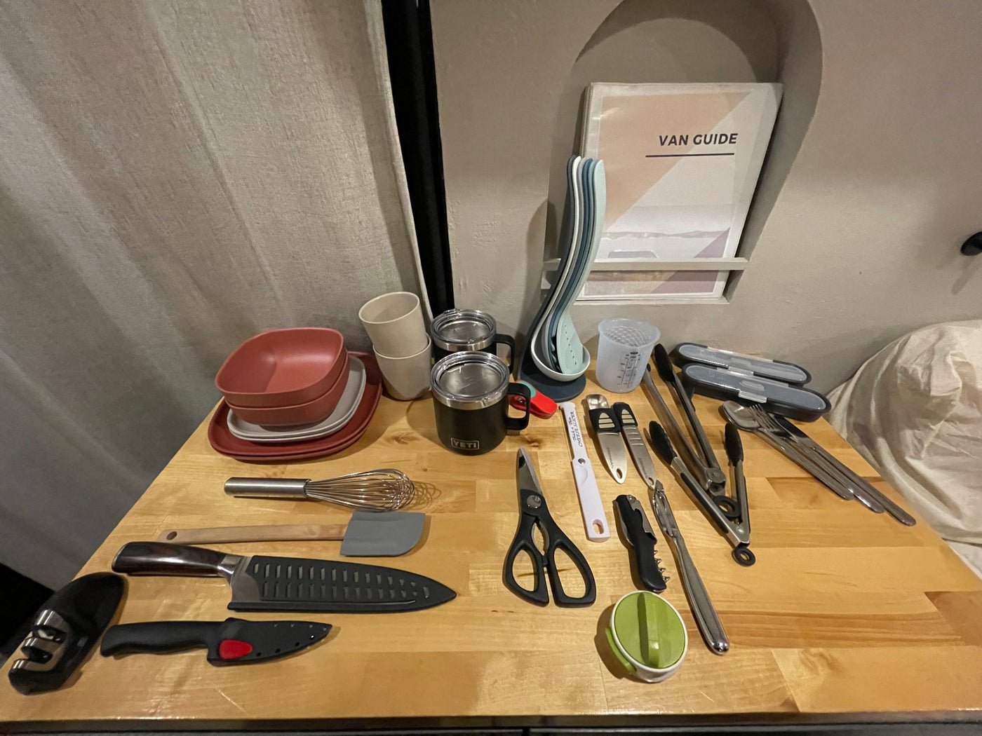 RV rental kitchen essentials: plates, cups, YETI mugs, knives, spatula, and various utensils on a wooden counter. — photo 13