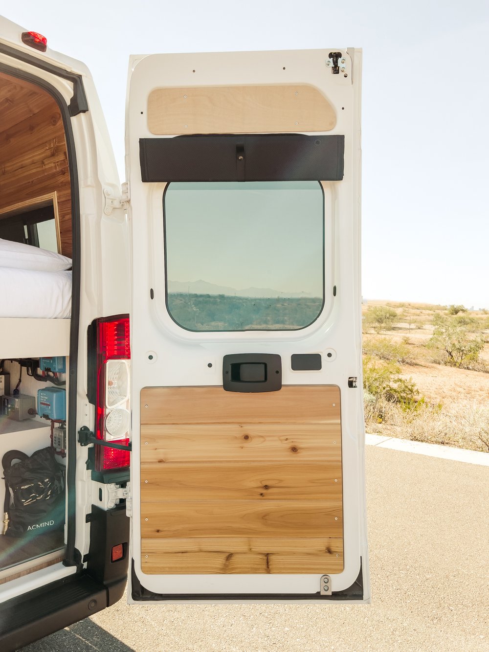 Inside an RV rental camper van, the open rear doors frame a bed, wood paneling, and a stunning desert landscape.