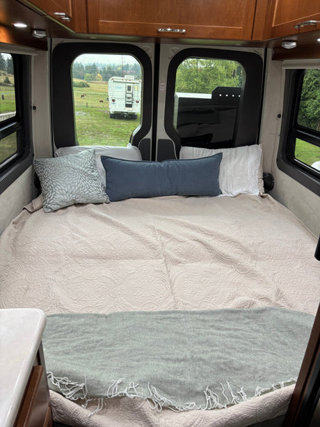 Cozy camper van bed for RV rental, featuring soft quilt, pillows, and serene views of a field and distant RV.