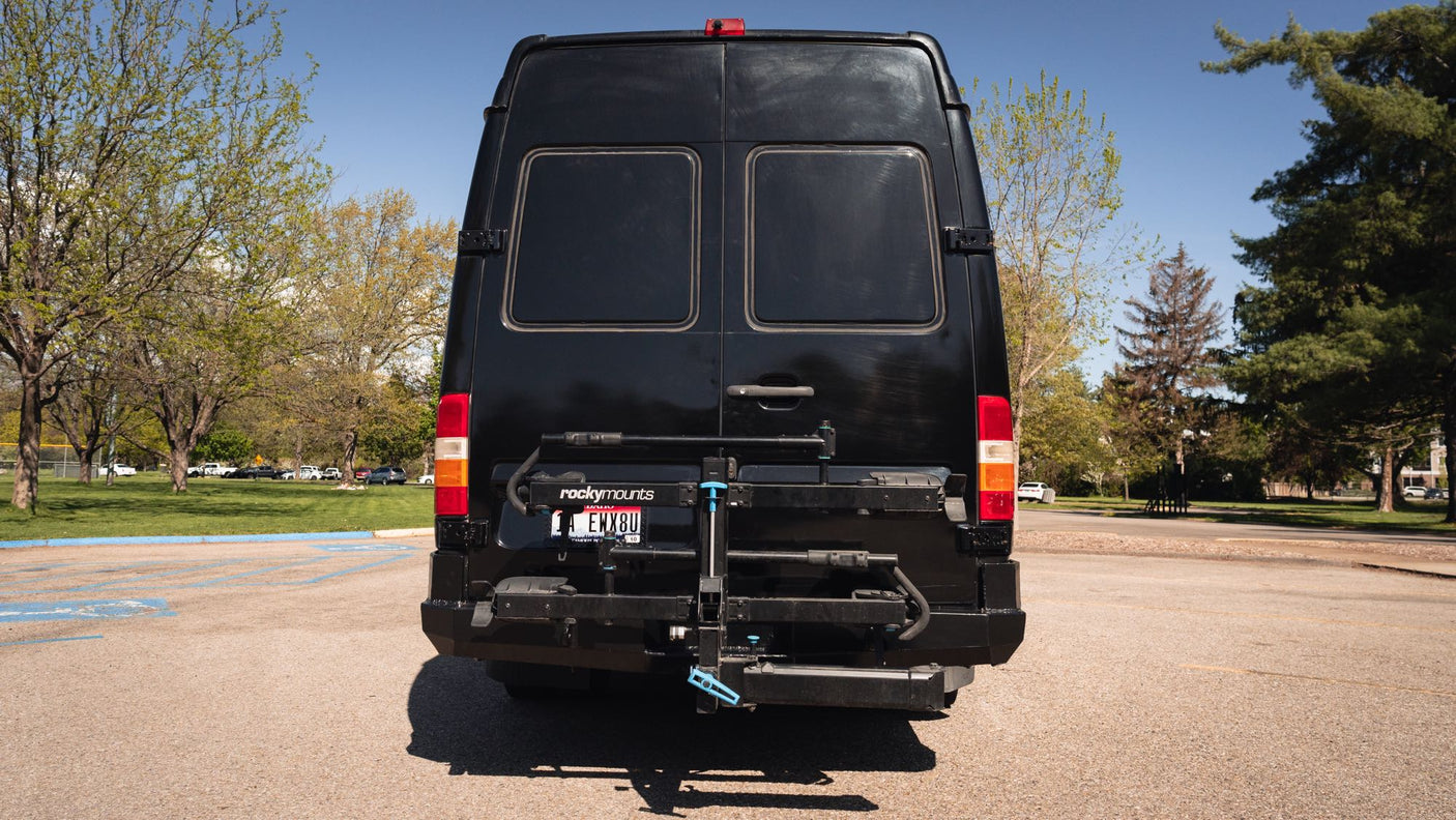 Black RV rental van with bike rack shown from behind, parked outside on a sunny day.
