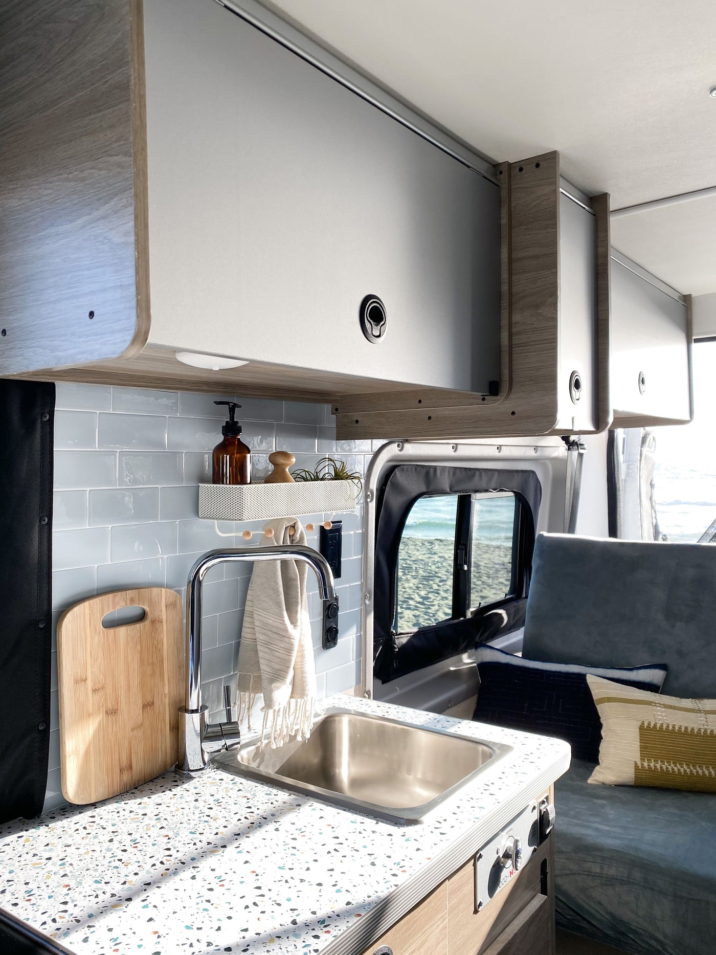 Inside an RV rental camper van: A modern kitchen area with sink, speckled counter, blue tile backsplash, & overhead storage. Beach view. — photo 3
