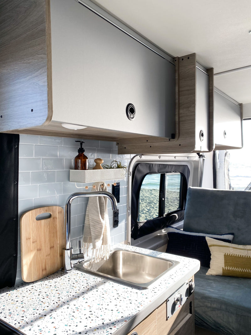 Inside an RV rental camper van: A modern kitchen area with sink, speckled counter, blue tile backsplash, & overhead storage. Beach view. — photo 3