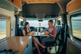Inside an RV rental, a couple enjoys a meal with a scenic lake view. Ideal for a comfortable van life dining experience.