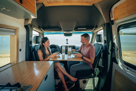 Inside an RV rental, a couple enjoys a meal with a scenic lake view. Ideal for a comfortable van life dining experience.