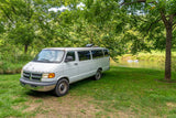 White van, suitable as an RV rental, parked on grass by a river with tubers & trees. Perfect for outdoor camping.