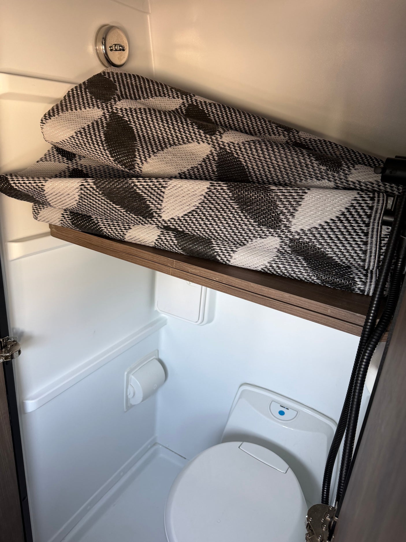 Compact RV rental bathroom features a toilet, shower hose, and a folded black and white mat on a shelf.