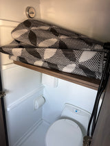 Compact RV rental bathroom features a toilet, shower hose, and a folded black and white mat on a shelf.