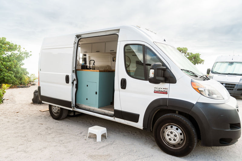White Ram Promaster camper van rv rental on sand with open side door showing teal & wood interior. Ideal for beach travel.|Bright RV rental interior features a functional kitchen with a sink — photo 1