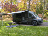Dark gray camper van with awning extended, chair & blanket on grass in autumn. Ideal for your RV rental getaway.