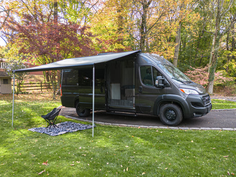 Dark gray camper van with awning extended, chair & blanket on grass in autumn. Ideal for your RV rental getaway.