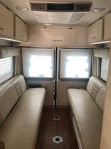 Inside this RV rental: a bright dinette area with opposing tan benches, wood floor, window blinds, and overhead storage.