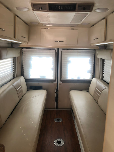 Inside this RV rental: a bright dinette area with opposing tan benches, wood floor, window blinds, and overhead storage.