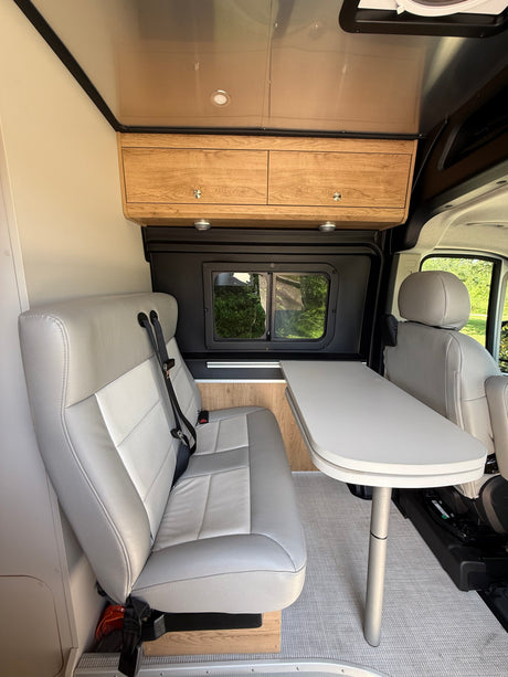 Modern RV rental interior with light gray seating, dining table, and wood cabinetry, offering a scenic window view.