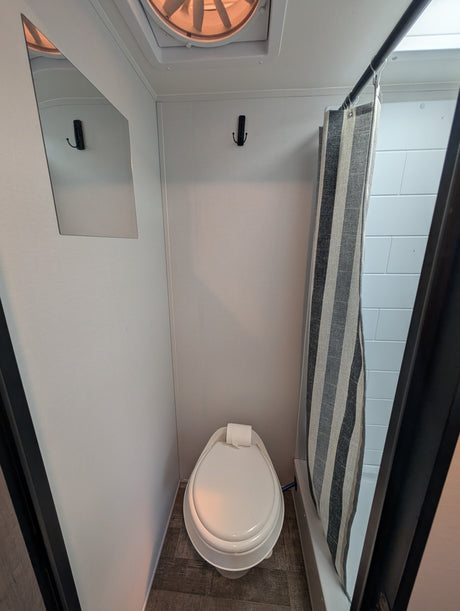 A compact RV rental wet bath with a toilet, striped shower curtain, wall mirror, and ceiling fan.