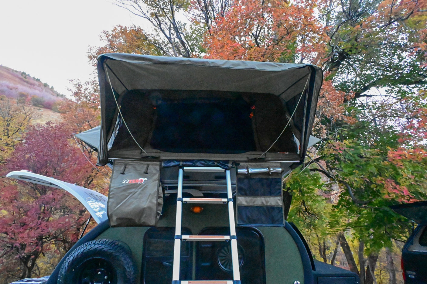 Enjoy autumn with this RV rental setup: a teardrop camper, rooftop tent & ladder amidst vibrant fall foliage. Great for camping!