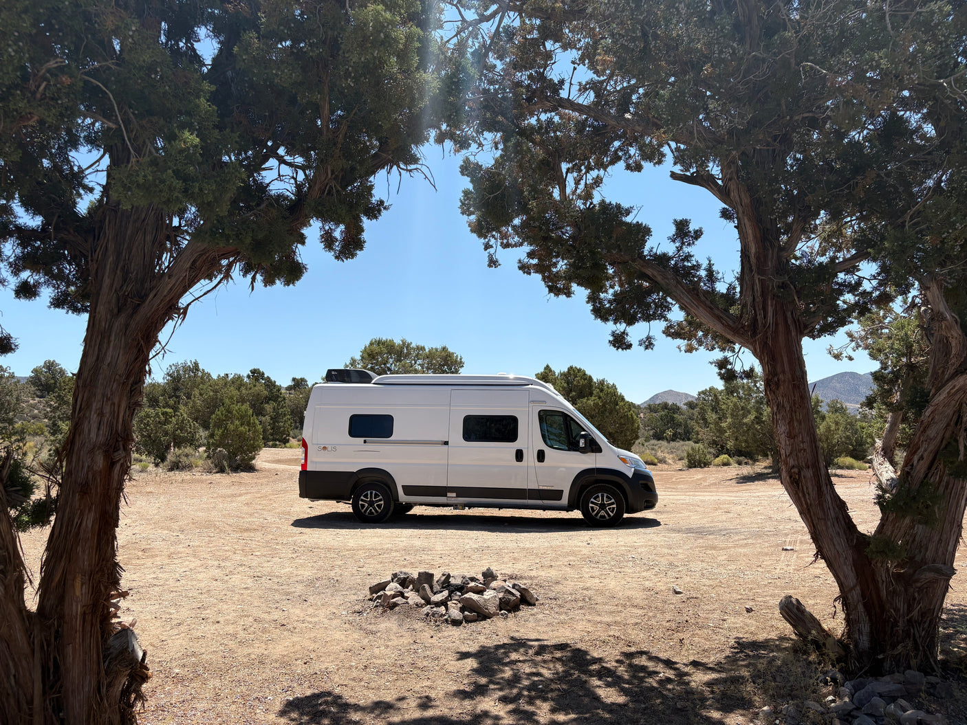 A white camper van, ideal as an rv rental, is parked in a dry, scenic landscape, framed by majestic trees.