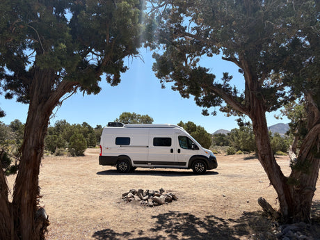 A white camper van, ideal as an rv rental, is parked in a dry, scenic landscape, framed by majestic trees.