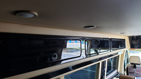 Inside an RV rental: view of custom black overhead cabinets, beige paneling, and ceiling lights. Spacious and modern.