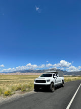 White Toyota Tundra pickup with an overland camper, reminiscent of an RV rental, parked on a scenic road with mountains and sand dunes.