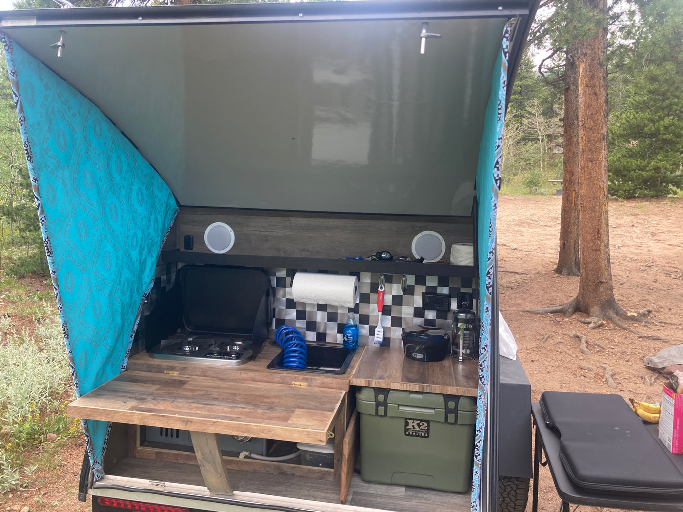 RV rental: Camper's open rear galley kitchen with stove, sink, and cooler, set up outdoors at a wooded campsite. — photo 3