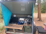 RV rental: Camper's open rear galley kitchen with stove, sink, and cooler, set up outdoors at a wooded campsite.
