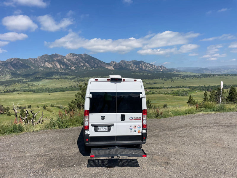  parked on gravel with scenic mountains and blue sky in background. A ramp extends from the rear.|Scenic RV rental: A white camper van parked on a dirt road with green valleys & distant mountains under a blue sky.|White RV rental van with deployed ramp — photo 4